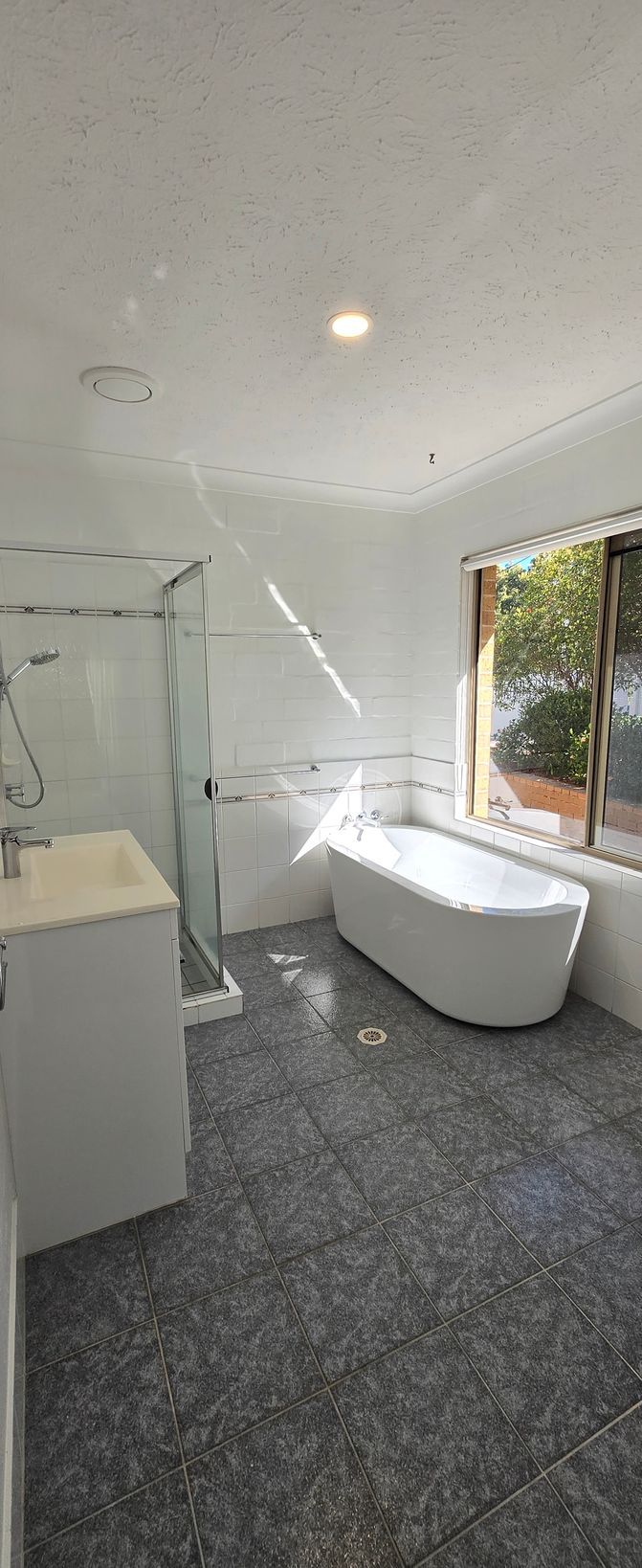 Bright bathroom with a freestanding tub, glass shower, gray tiles, and window light.