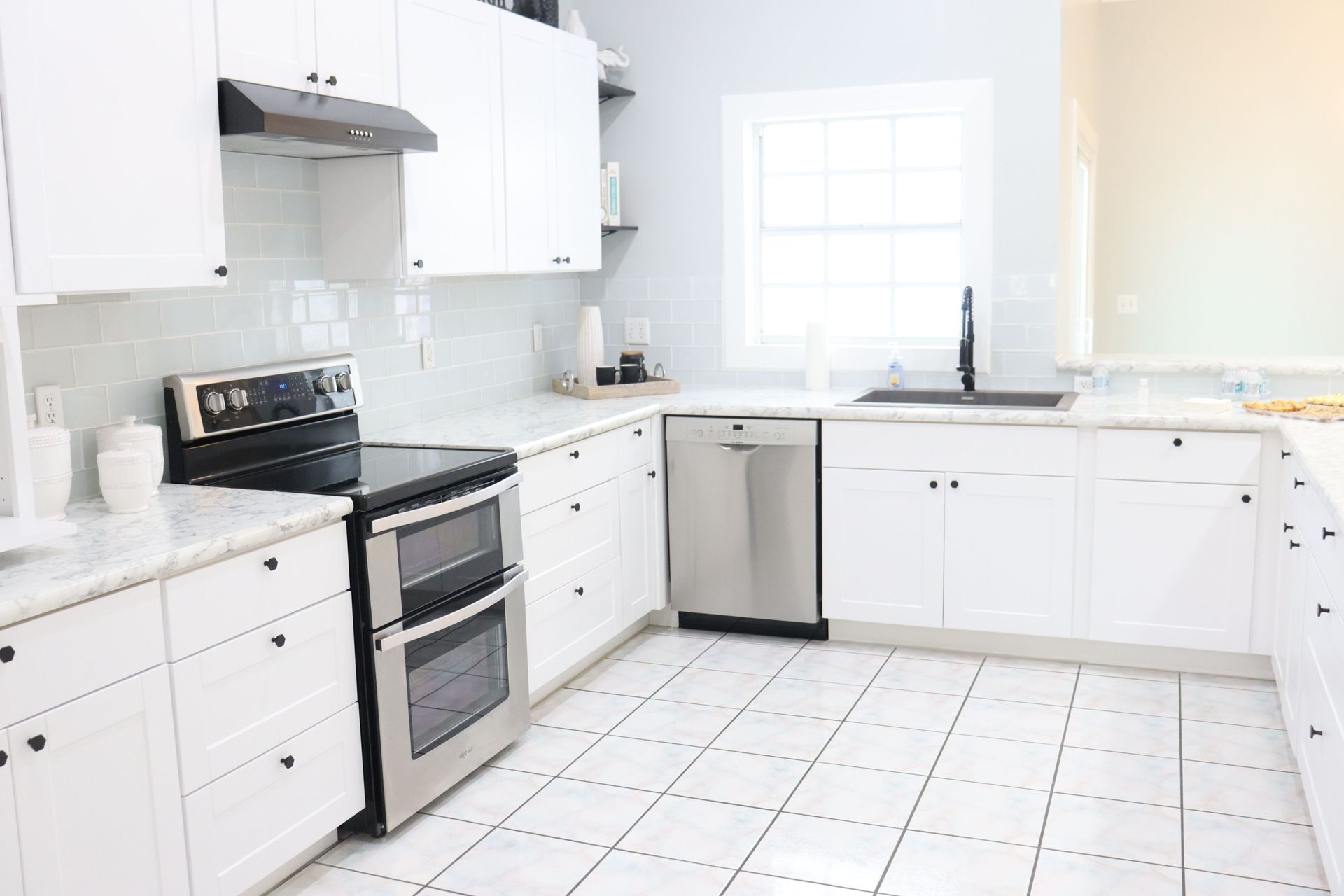 Bright white kitchen with stainless steel appliances, tiled floor, and a window over the sink