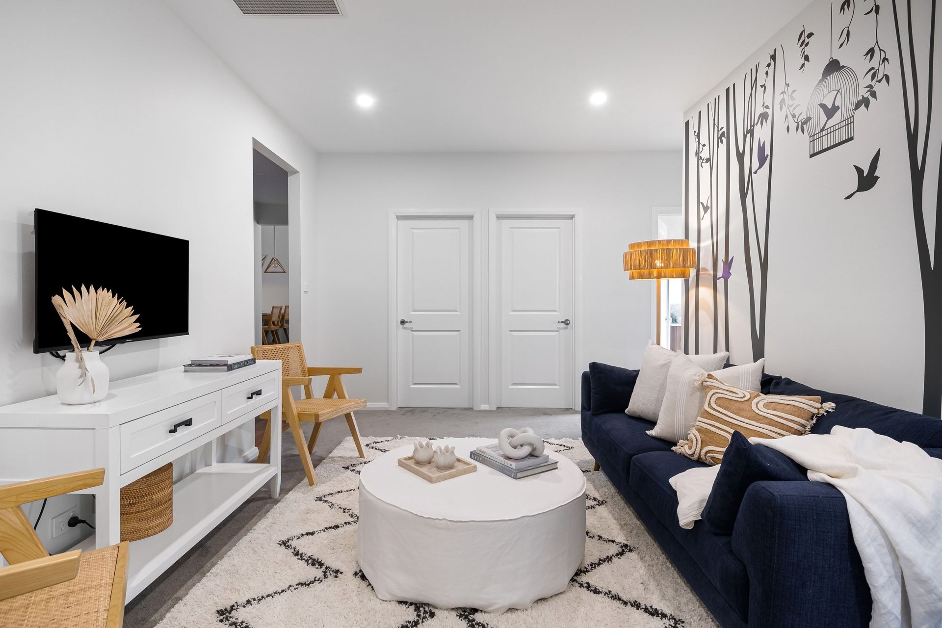 Bright white living room with navy sofa, round ottoman, TV console, and double doors.