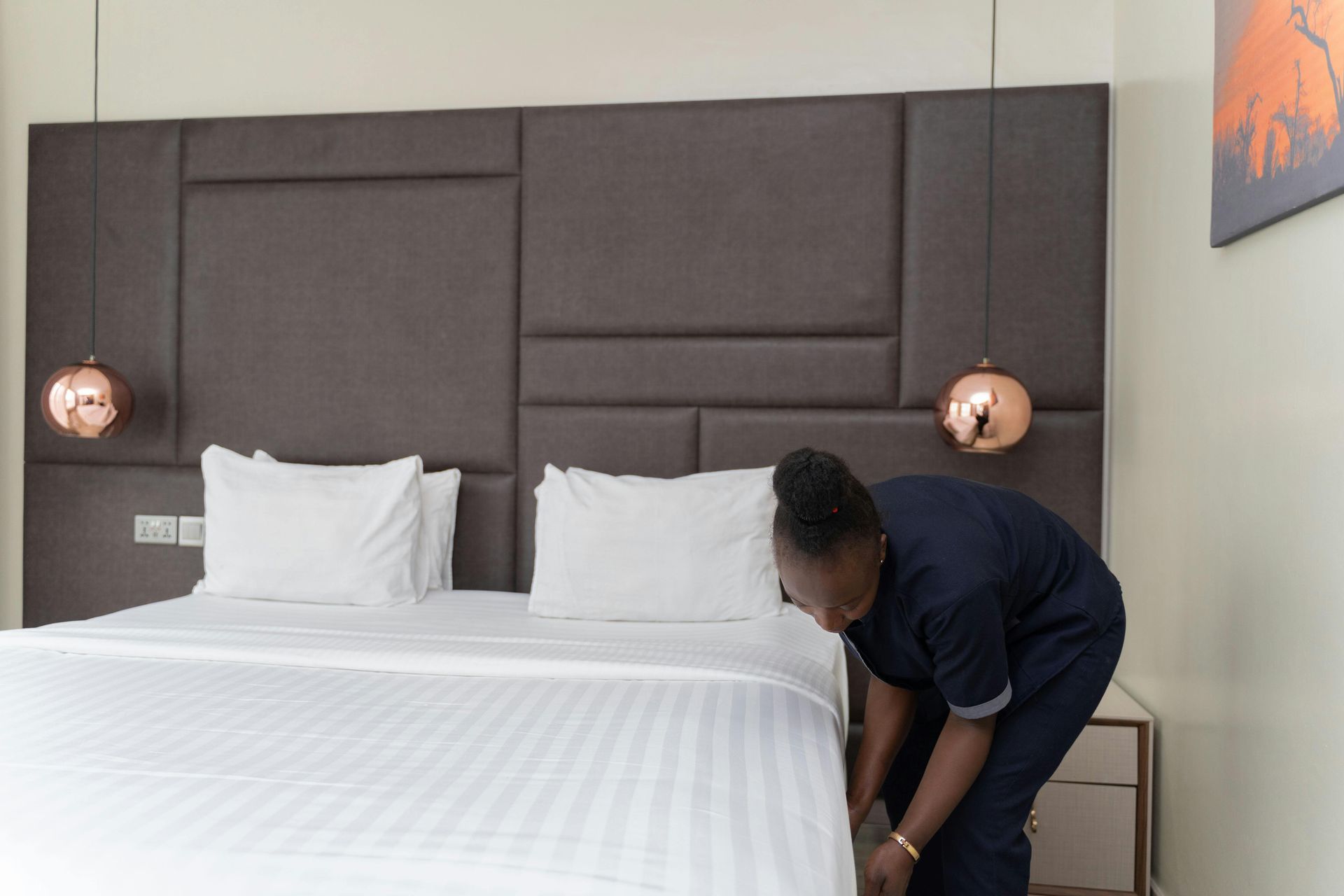 Person making a bed in a modern hotel room with white linens and gray wall panels