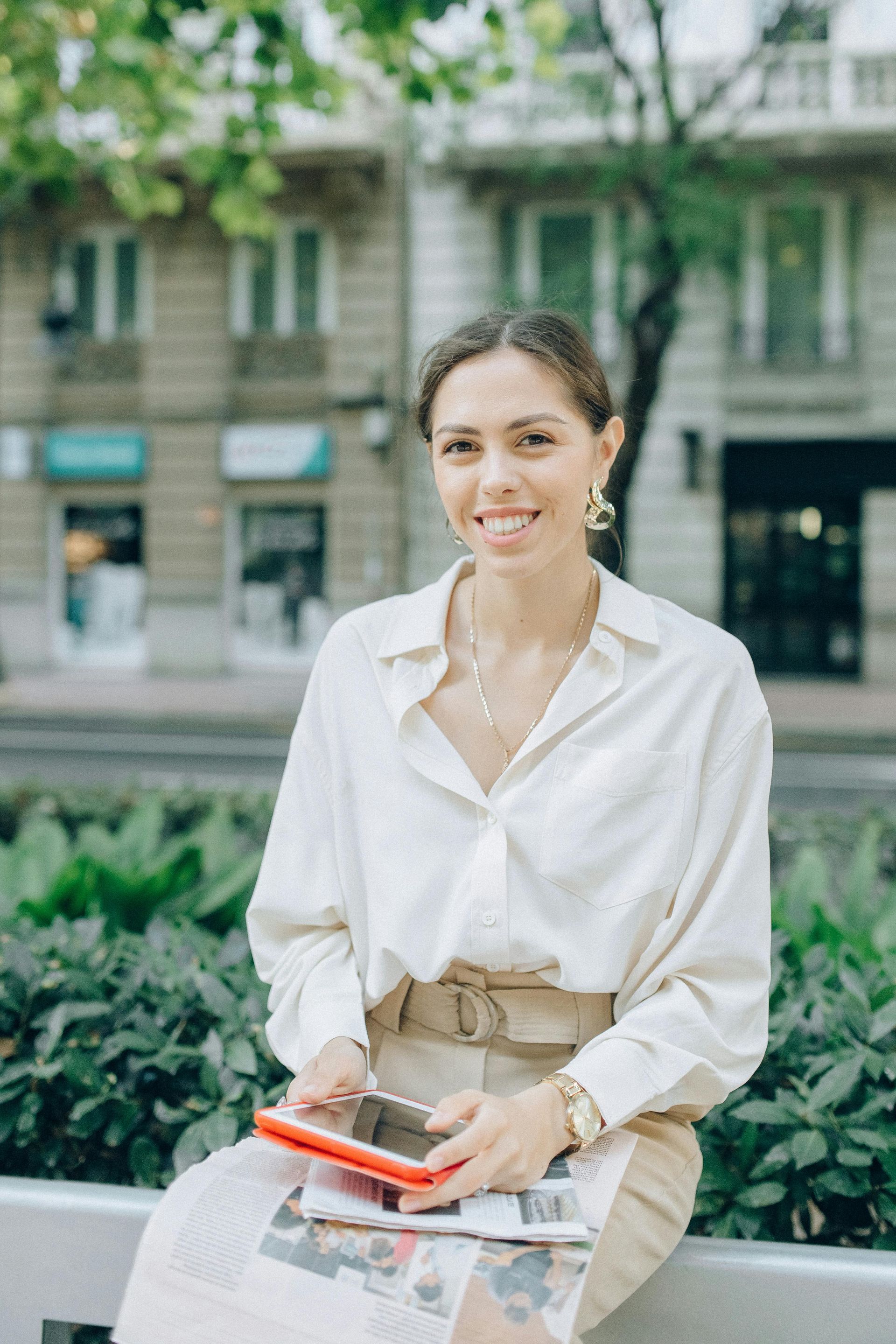 Smiling woman in a white blouse and beige skirt holding a notebook on a city bench.