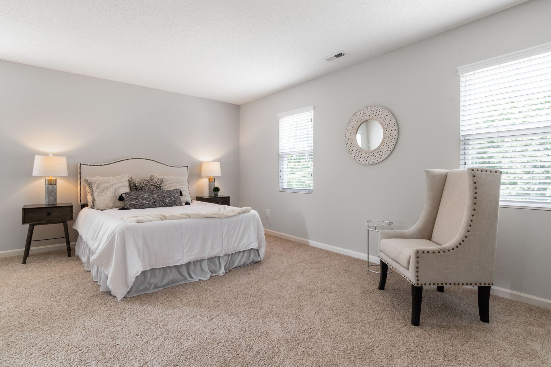 Spacious bedroom with a bed, two lamps, a chair, and a wall clock on light gray walls.
