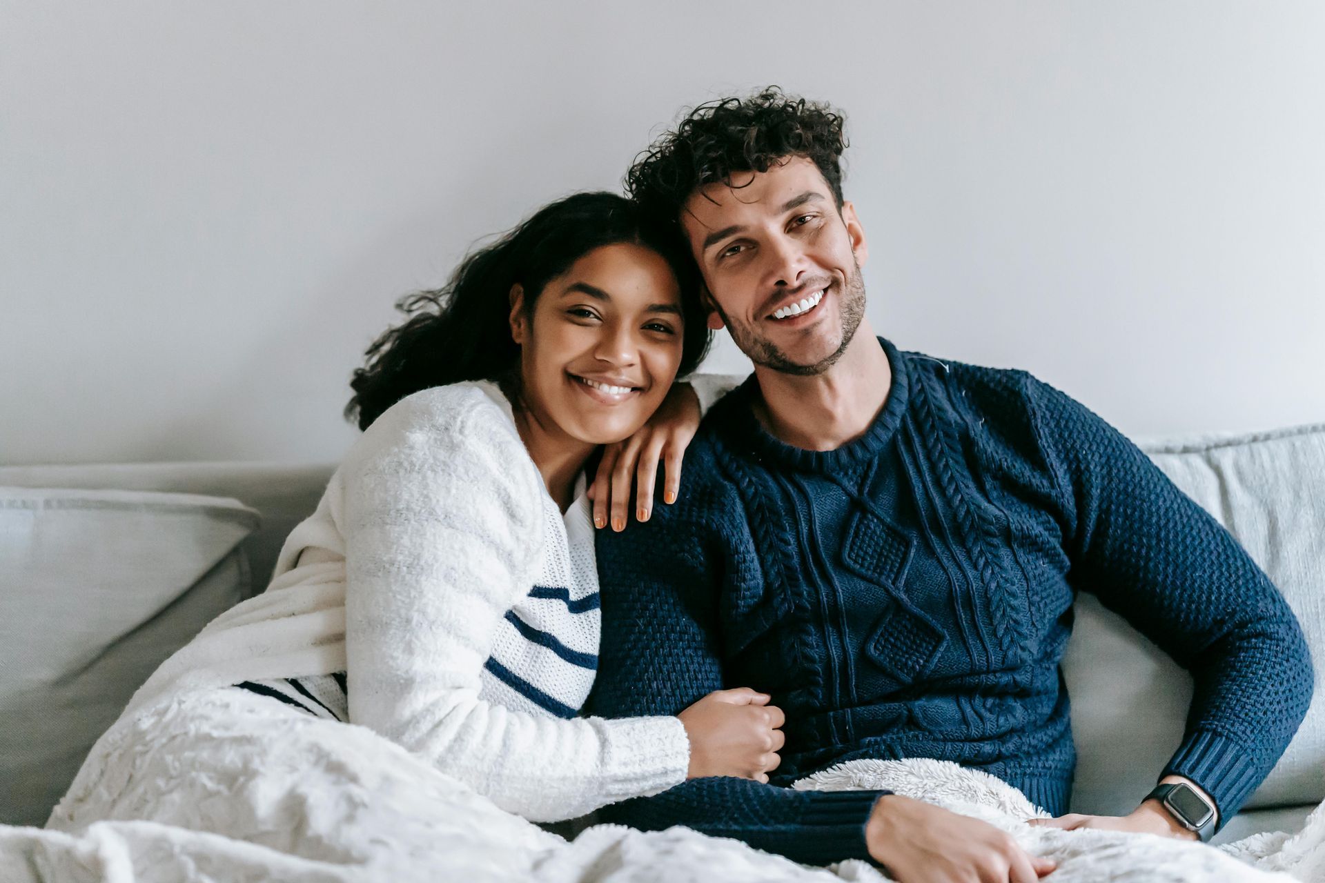 Couple smiling on a couch under a gray blanket, wearing white and navy sweaters
