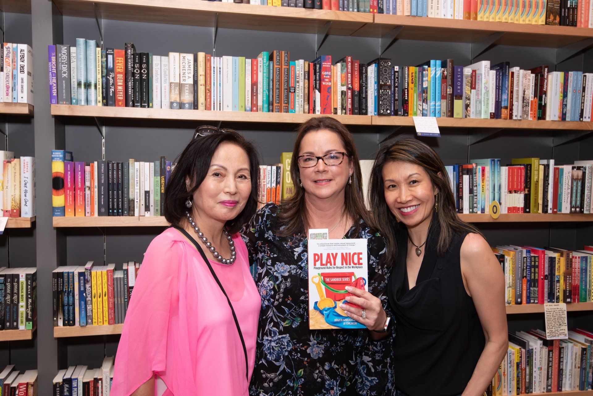Three women are standing in front of a bookshelf holding a book titled play mice.