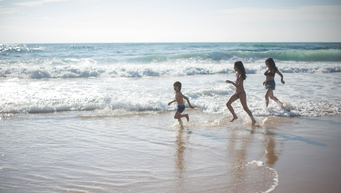 Three people running in shallow water towards ocean waves on a sunny beach.