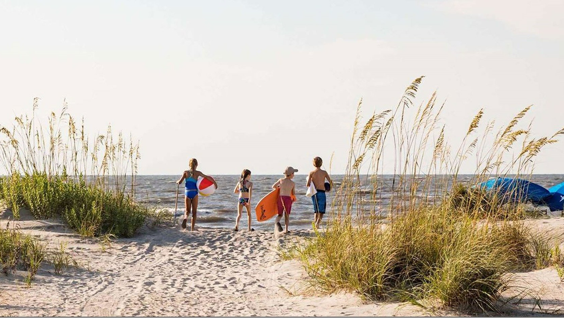 People walking towards the ocean, carrying boogie boards, beach grass in foreground.