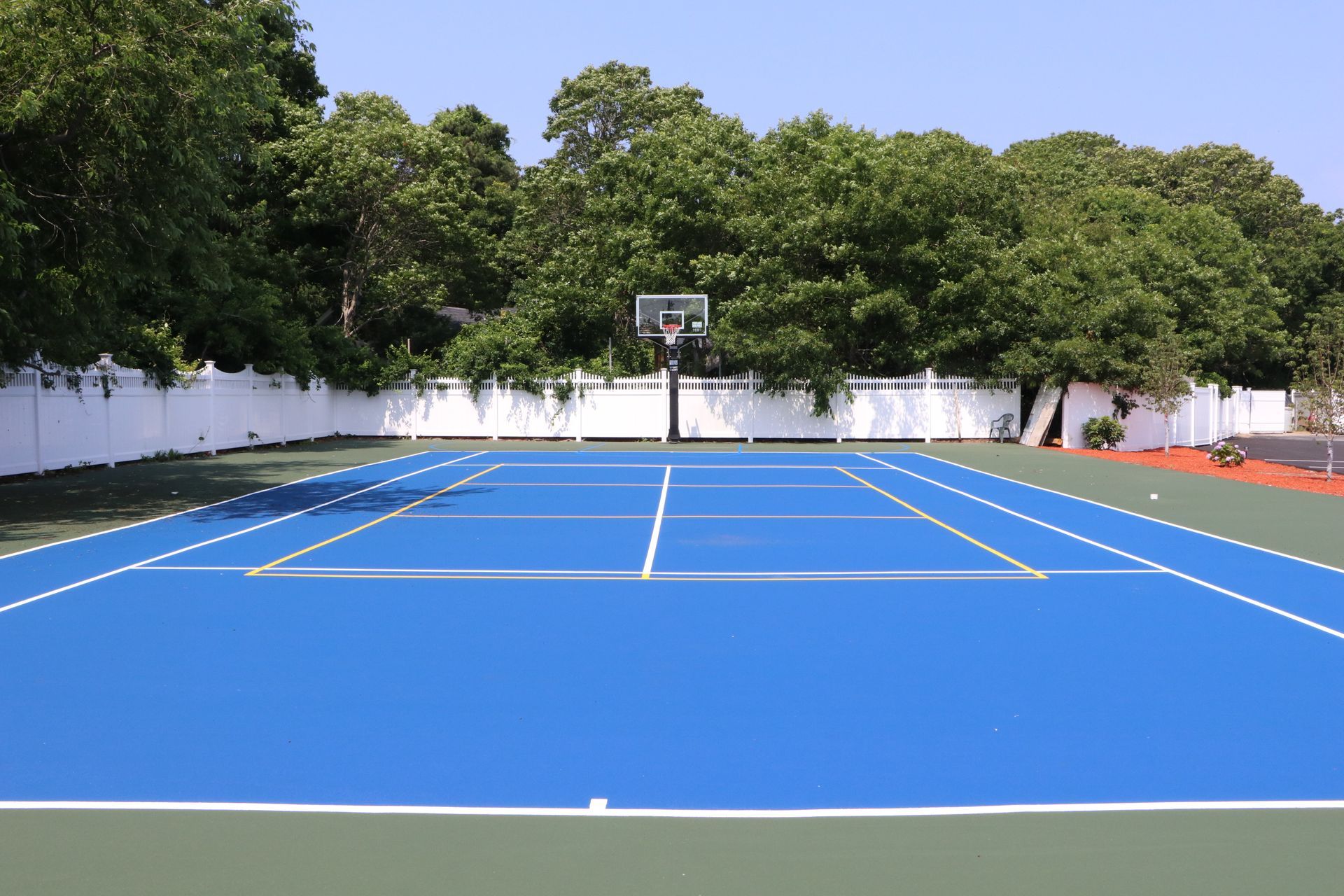 Blue and green tennis court with white lines, basketball hoop, and white fence.