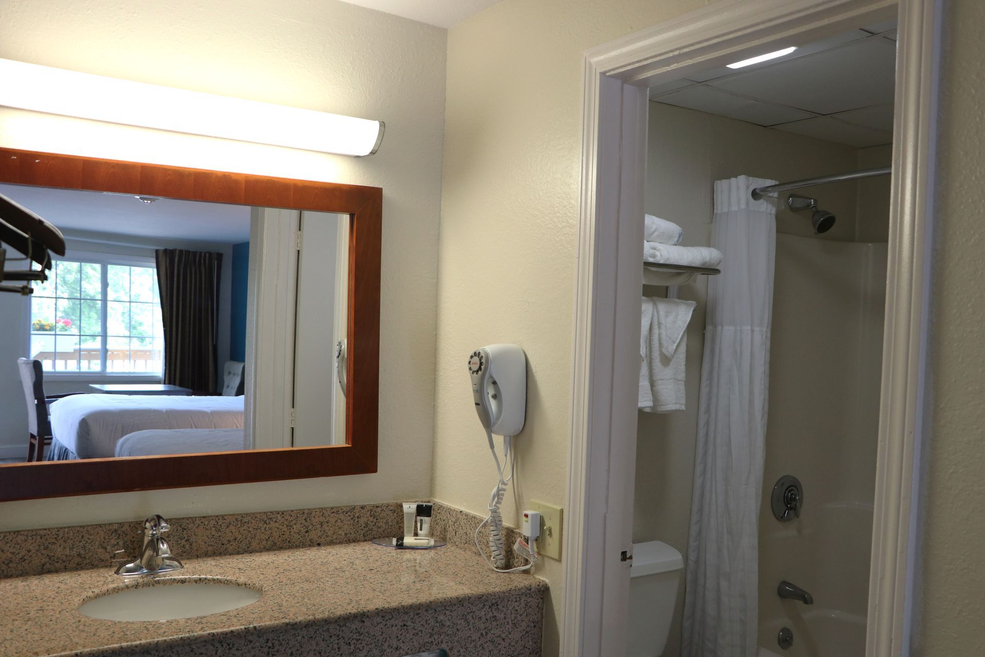 Bathroom with mirror reflecting a hotel bedroom, granite countertop, white shower curtain, and hand dryer.