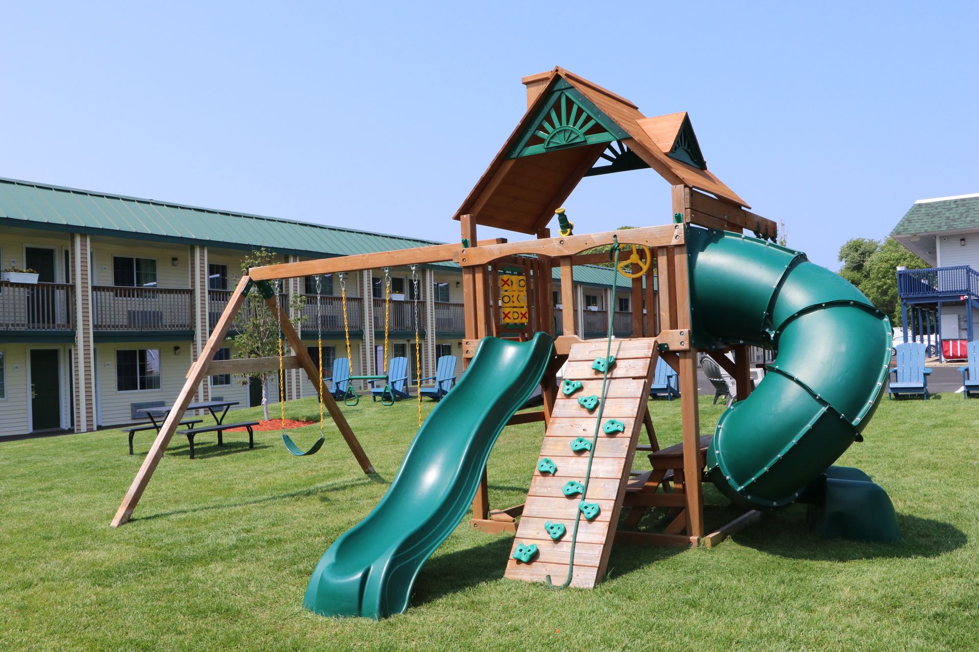 Playground with green slide, climbing wall, and swings in front of a motel building.