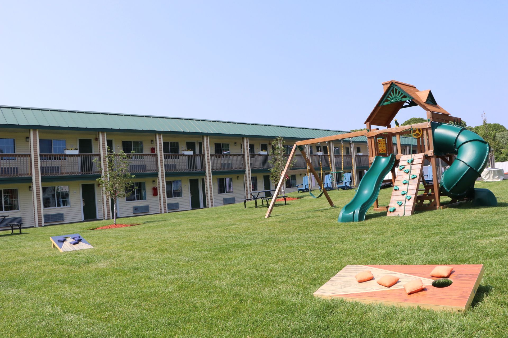 Motel exterior with a playground on a grassy hill; a swing set, slide, climbing wall, and cornhole game.