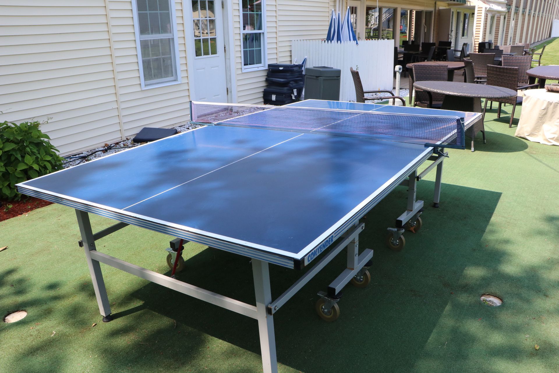 Outdoor blue ping pong table on a green surface, near a building and patio furniture.