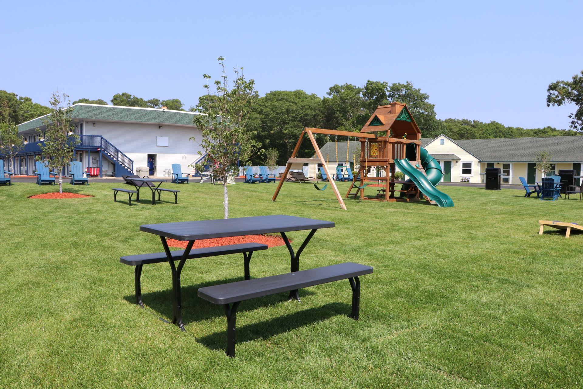 A grassy outdoor area with picnic table, playground, and motel units on a sunny day.