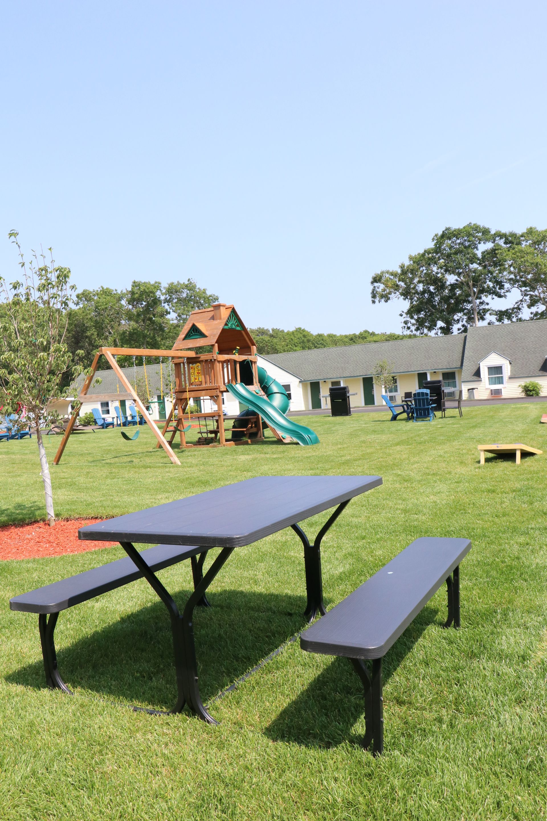 Picnic table with benches in a grassy yard, with a playground in the background and a blue sky.