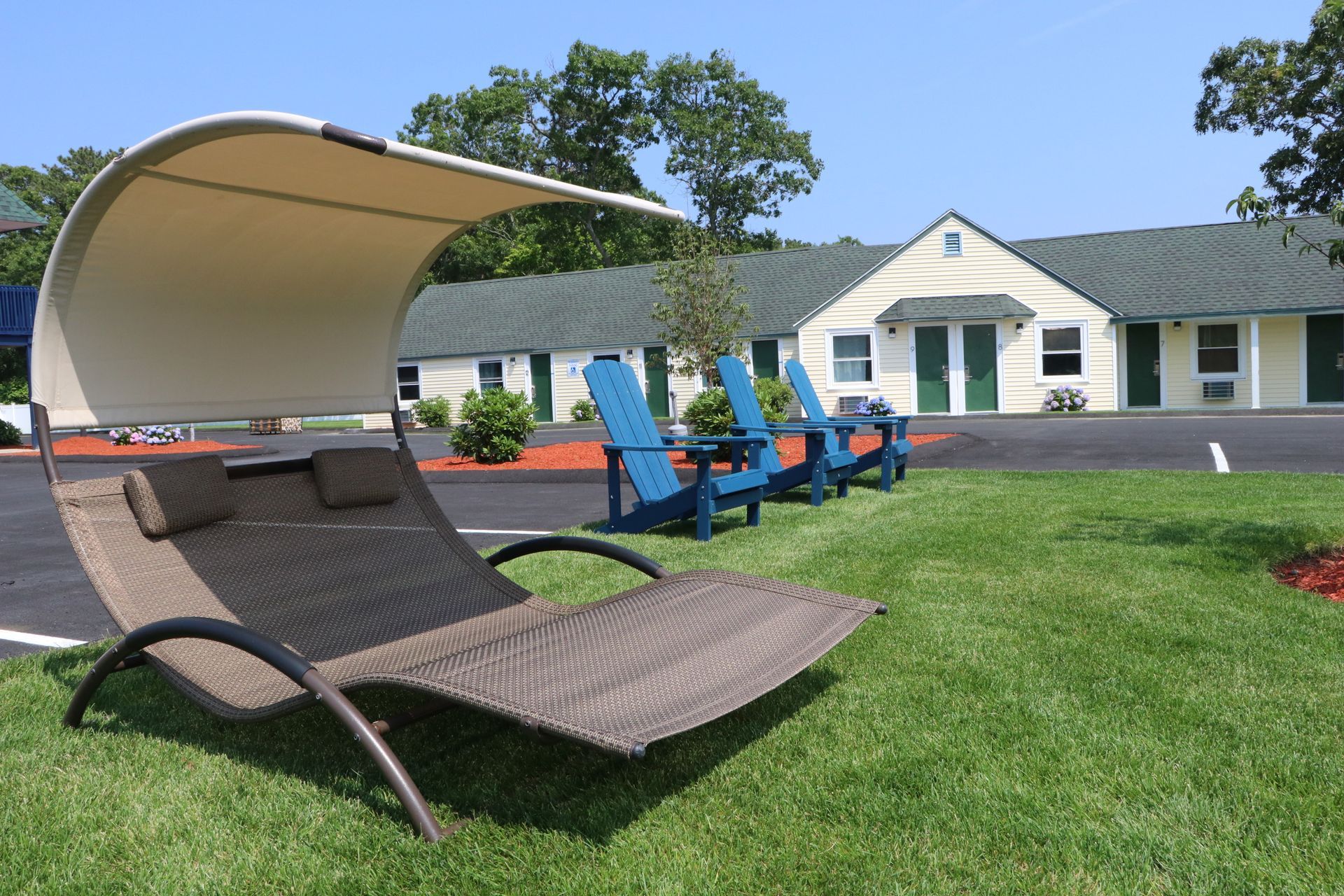 Lounge chair with canopy on a lawn, blue Adirondack chairs in background, motel exterior. Sunny day.