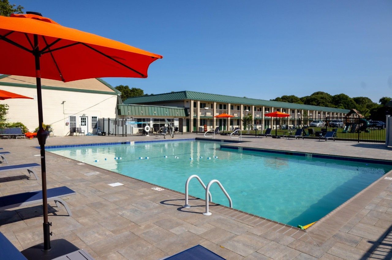 A rectangular pool with clear blue water, surrounded by gray paving stones, orange umbrellas, and a two-story motel.