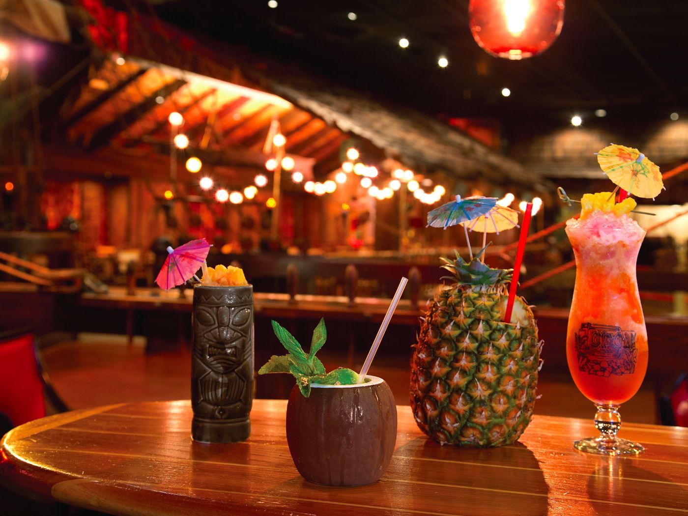 Cocktails on bar in tiki bar setting: tiki mug, coconut, pineapple, and tall glass with umbrellas and fruit garnishes.