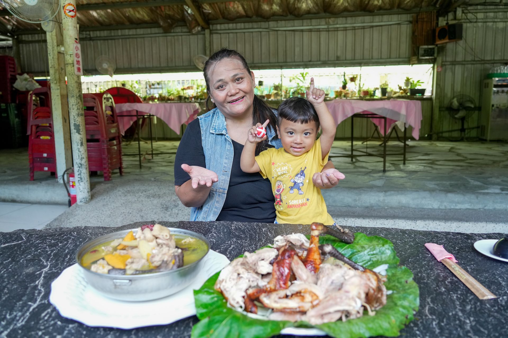 一位茂林部落婦女帶著孩子坐在擺有中興紅羽1982土雞製作的美味菜餚的餐桌旁。孩子舉起了一隻手。他們正在一家露天餐廳用餐。