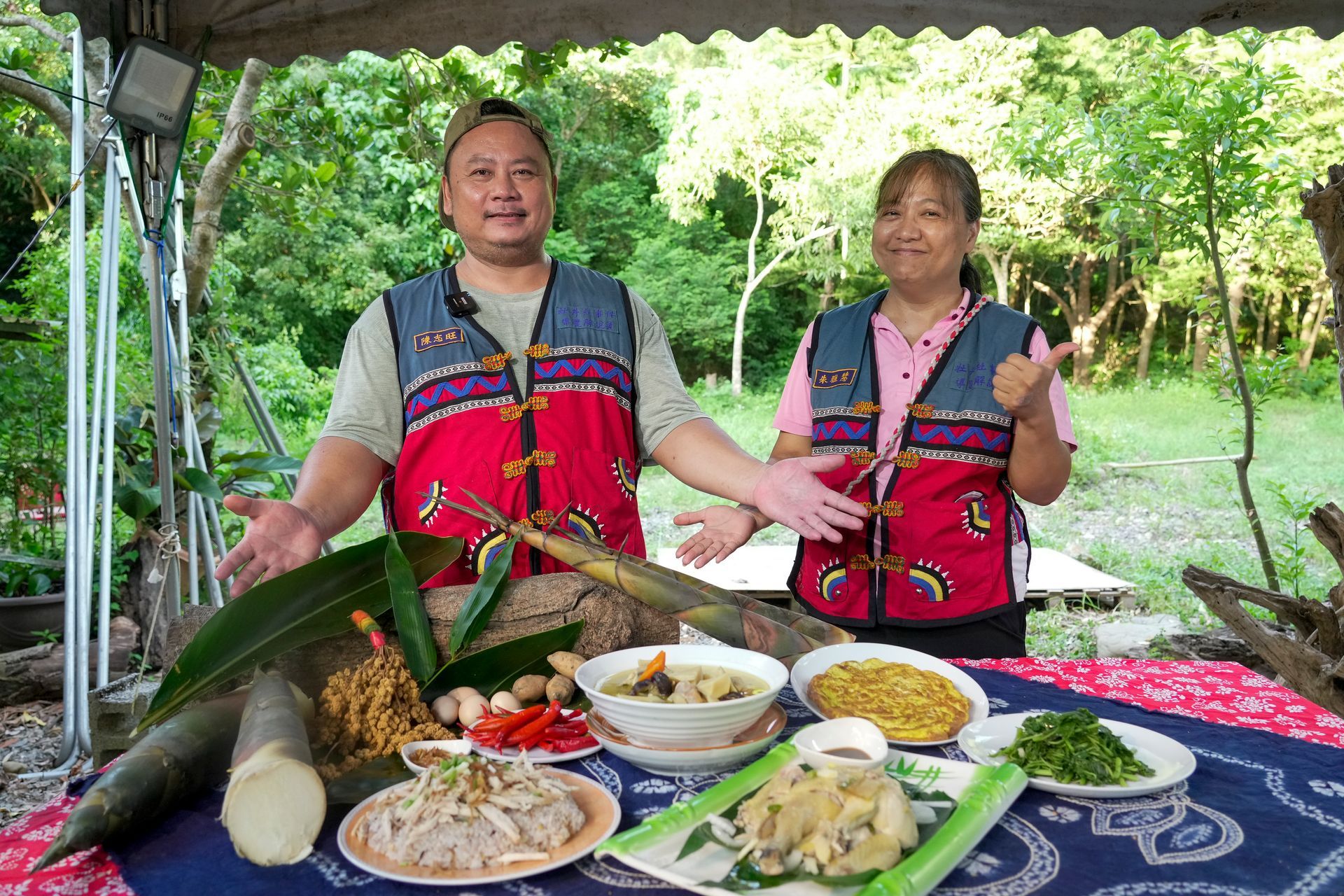 兩個人站在擺滿食物的桌子後面，面帶微笑。桌子周圍擺放著中興紅羽1982土雞做的熟食、竹子和綠葉。
