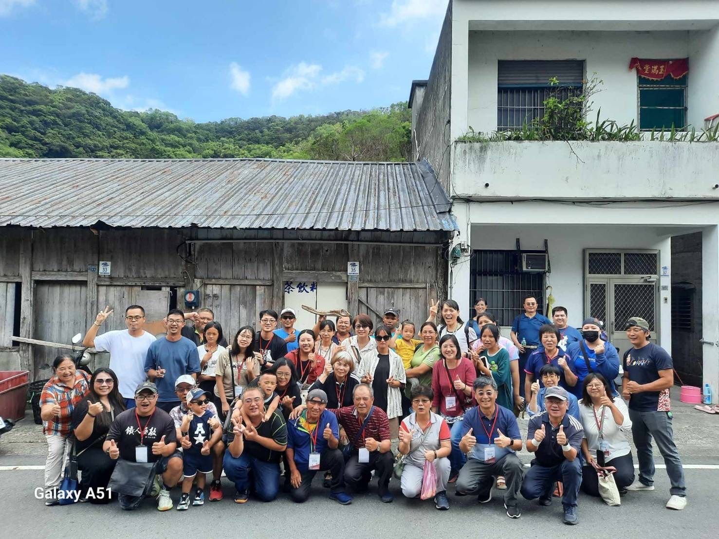 一群人在戶外建築附近擺姿勢拍照,背景是山。許多人面帶微笑。