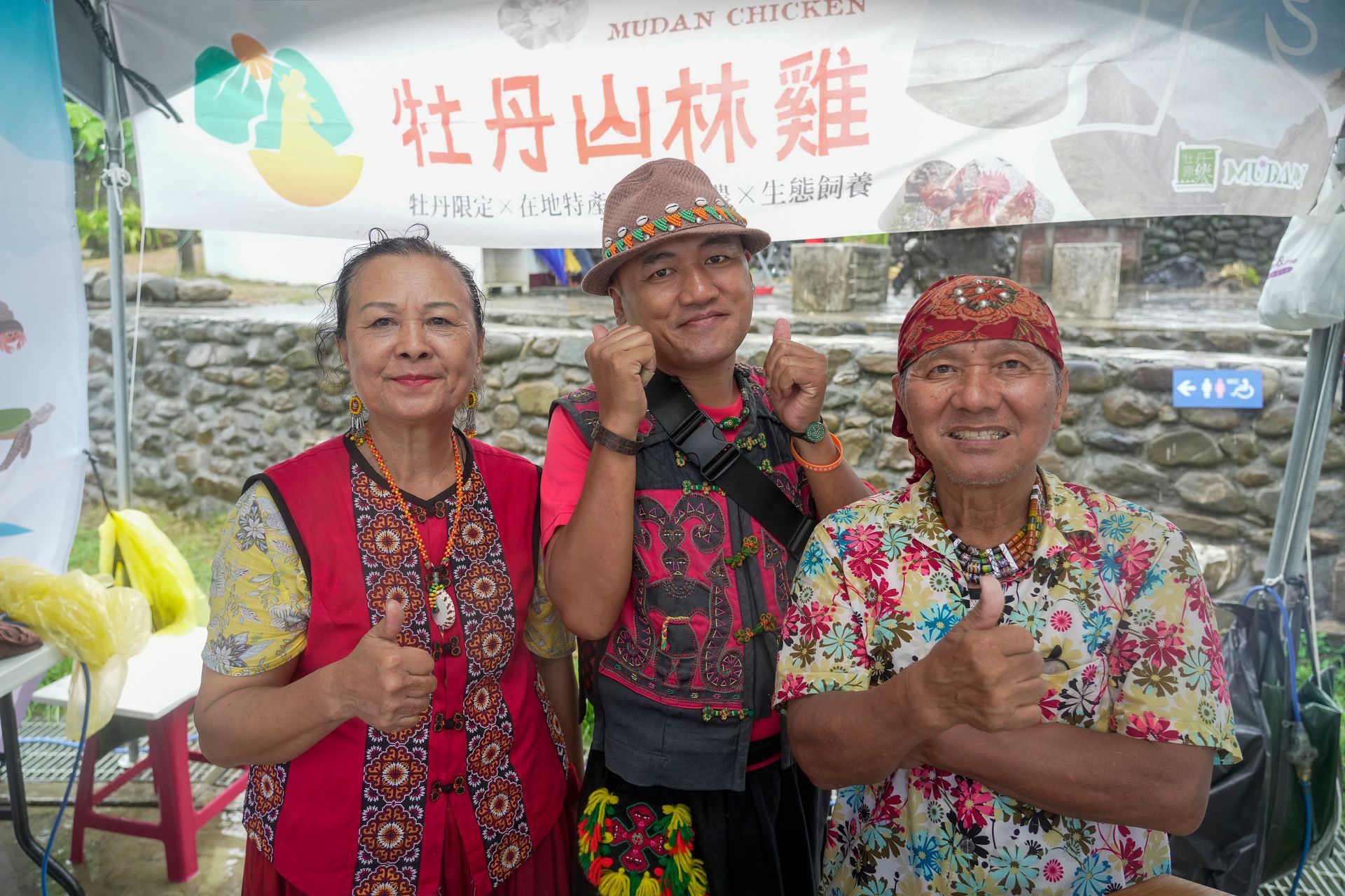 Three smiling people give thumbs up at an outdoor food stall. A banner reads 