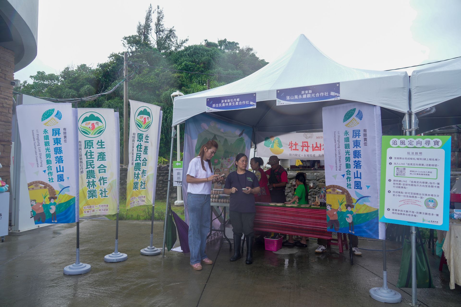 牡丹鄉部落戶外市場牡丹山林雞的攤位上,兩位女士正在交談。白色帳篷裡掛著旗幟,擺放著商品。陰天。