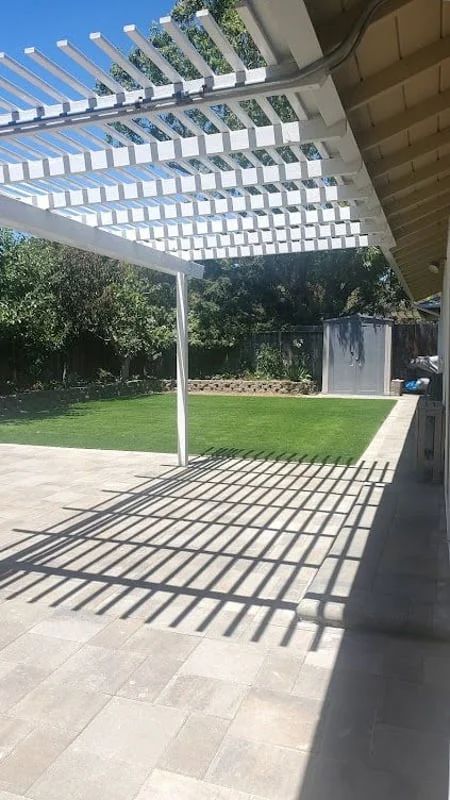 Patio with white pergola casting striped shadows on a paved walkway beside a green lawn.