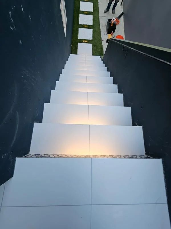Outdoor white staircase descending between dark walls, with a grassy landing and people at the top.