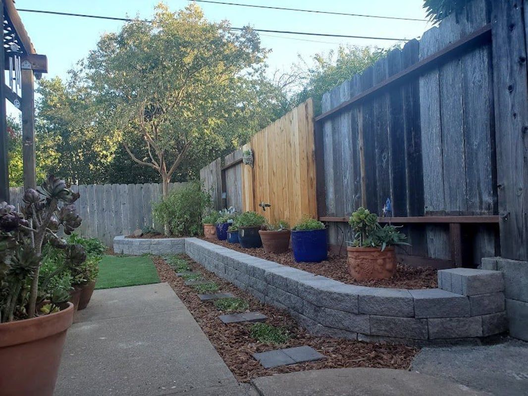 Backyard with concrete path, raised garden bed, wooden fence, and potted plants.