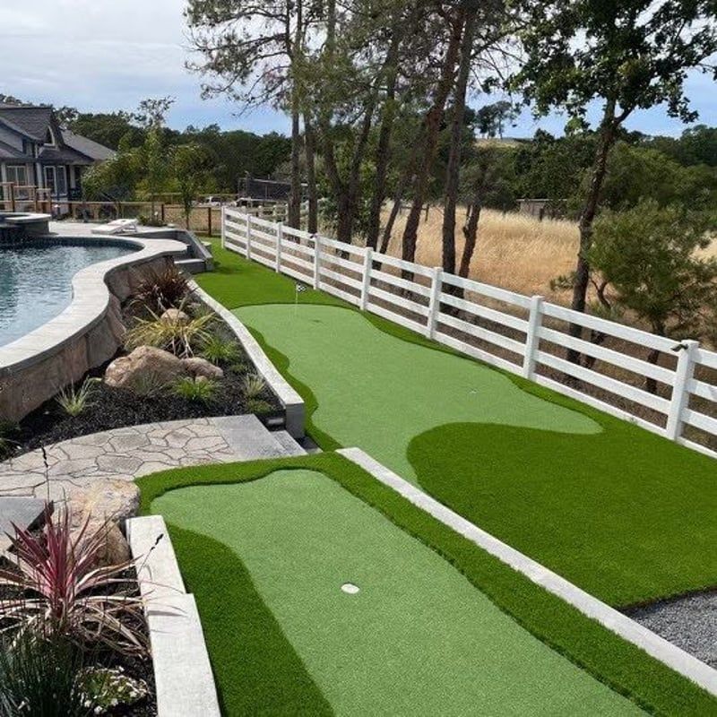 Backyard putting green beside a pool, with white fence and trees on a sunny day