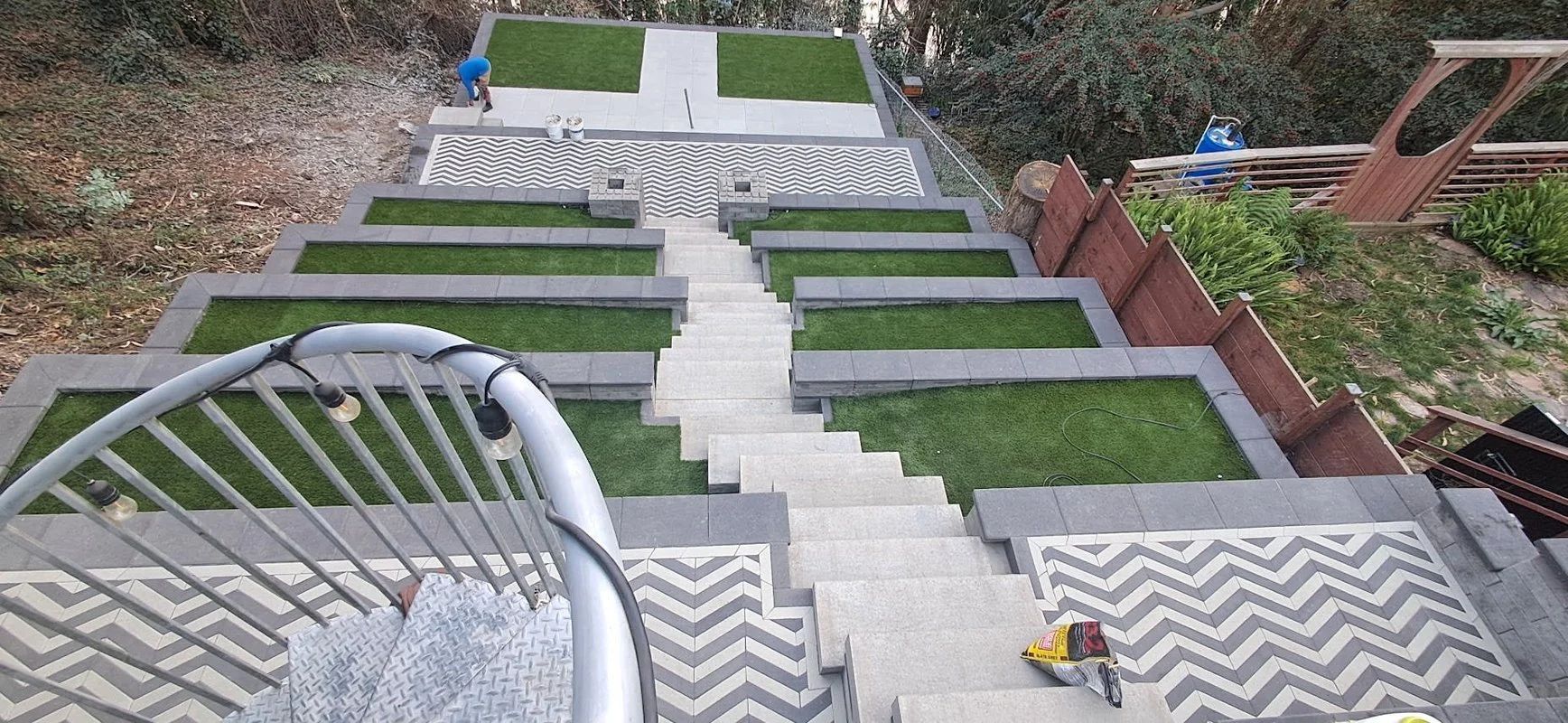 Steep outdoor staircase with gray zigzag steps, white walls, green grass terraces, and a metal handrail