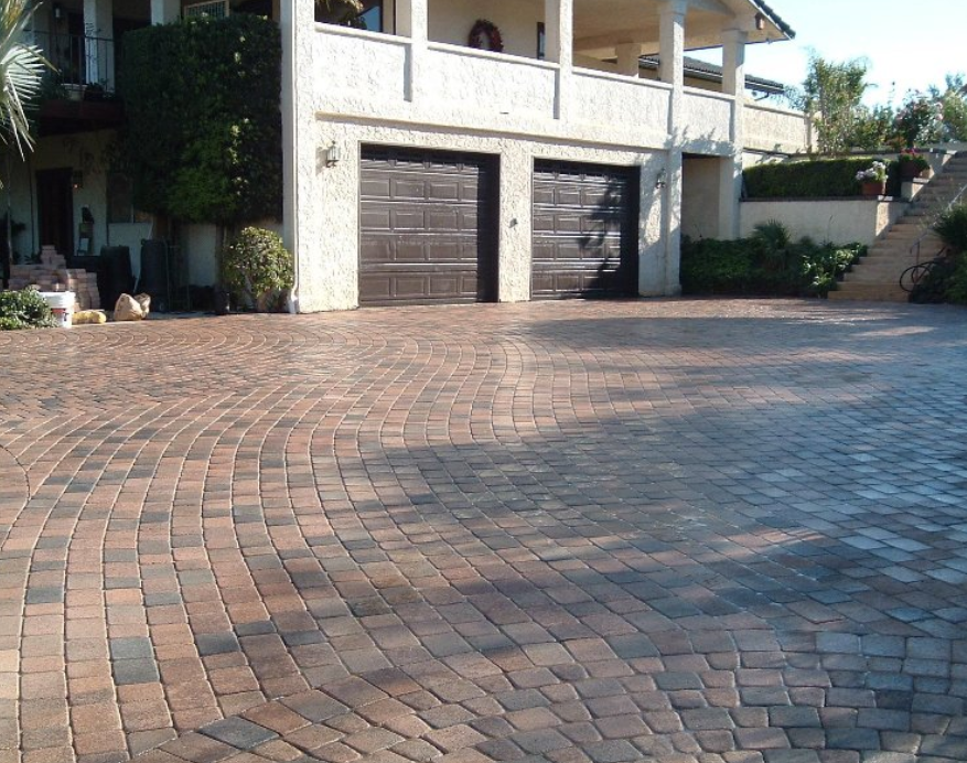 Brick-paved driveway leading to a house with two dark garage doors and stucco exterior Brick-paved driveway leading to a house with two dark garage doors and stucco exterior