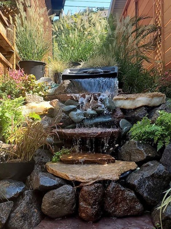 Small backyard stone waterfall with flowing water, surrounded by rocks, plants, and a narrow garden path