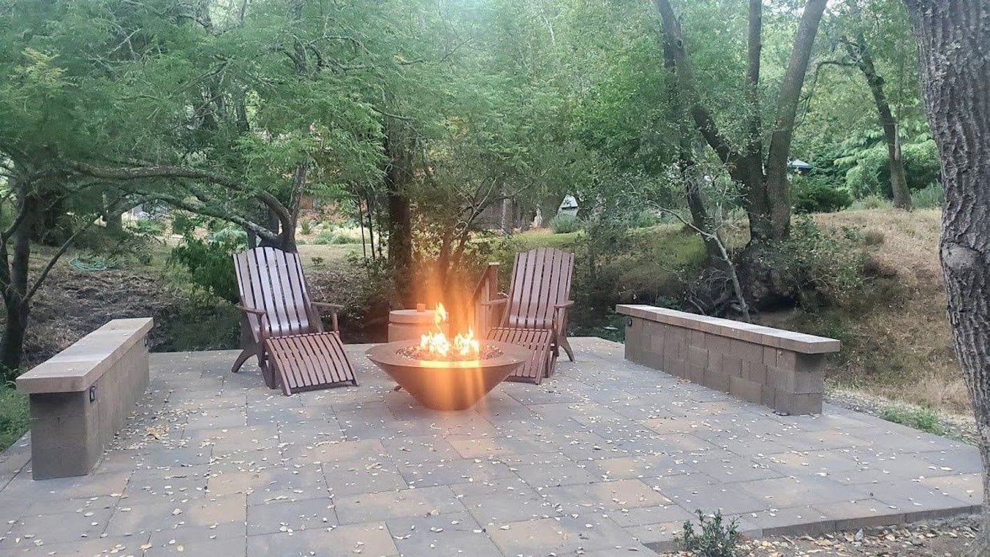 Wooded campsite with a small fire pit, two chairs, and stone benches on a concrete pad