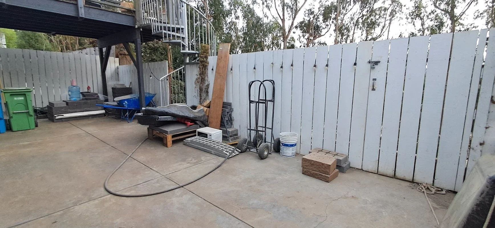 Backyard patio with scattered items, a wheelbarrow, bricks, and a white wooden fence under trees