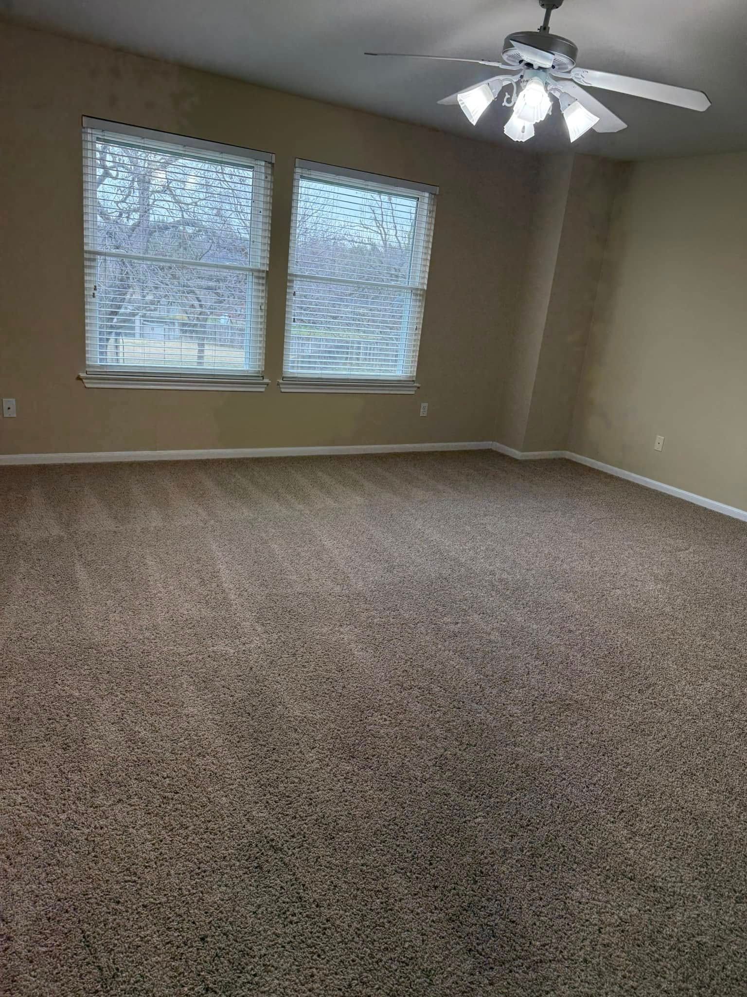 An empty living room with a ceiling fan and two windows.