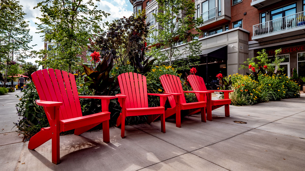 A row of red adirondack chairs are sitting on a sidewalk in front of a building.
