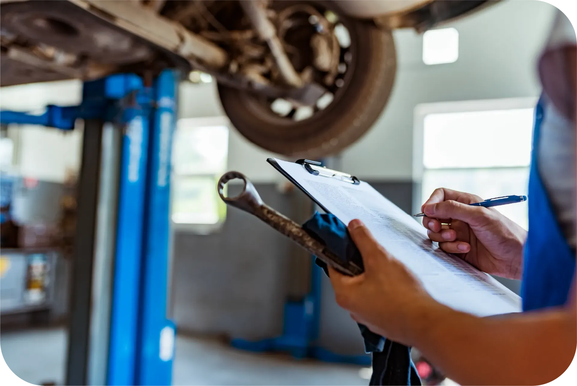 A mechanic is writing on a clipboard in front of a car on a lift.