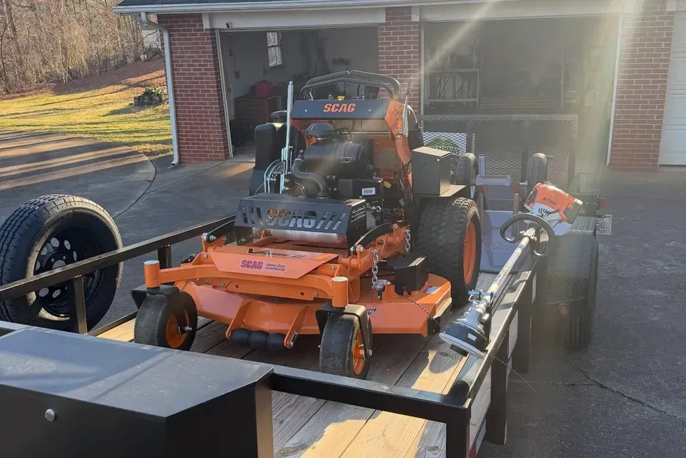 Orange riding lawn mower on a trailer in a driveway beside a brick garage.