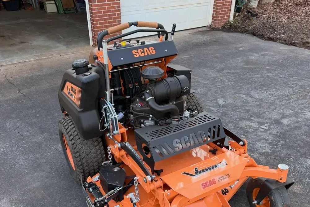 Orange SCAG lawn mower parked on a driveway beside a brick garage
