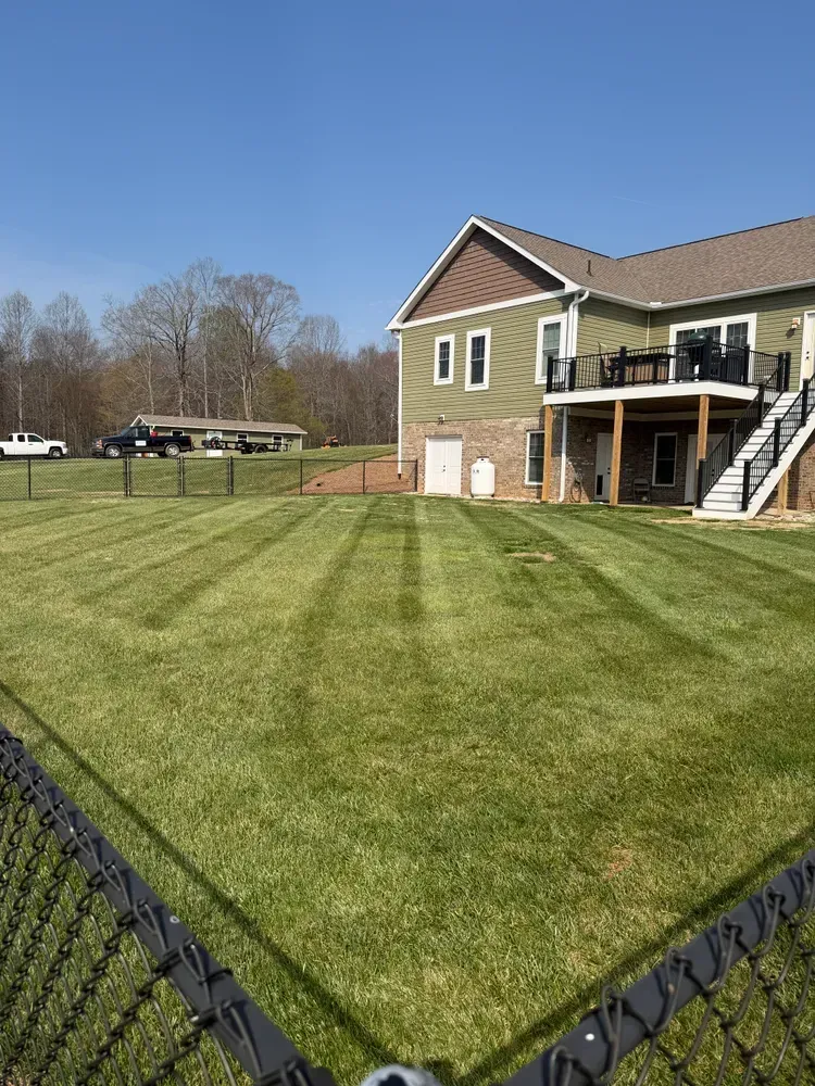Green striped lawn beside a brick house with stairs and a fenced yard on a sunny day