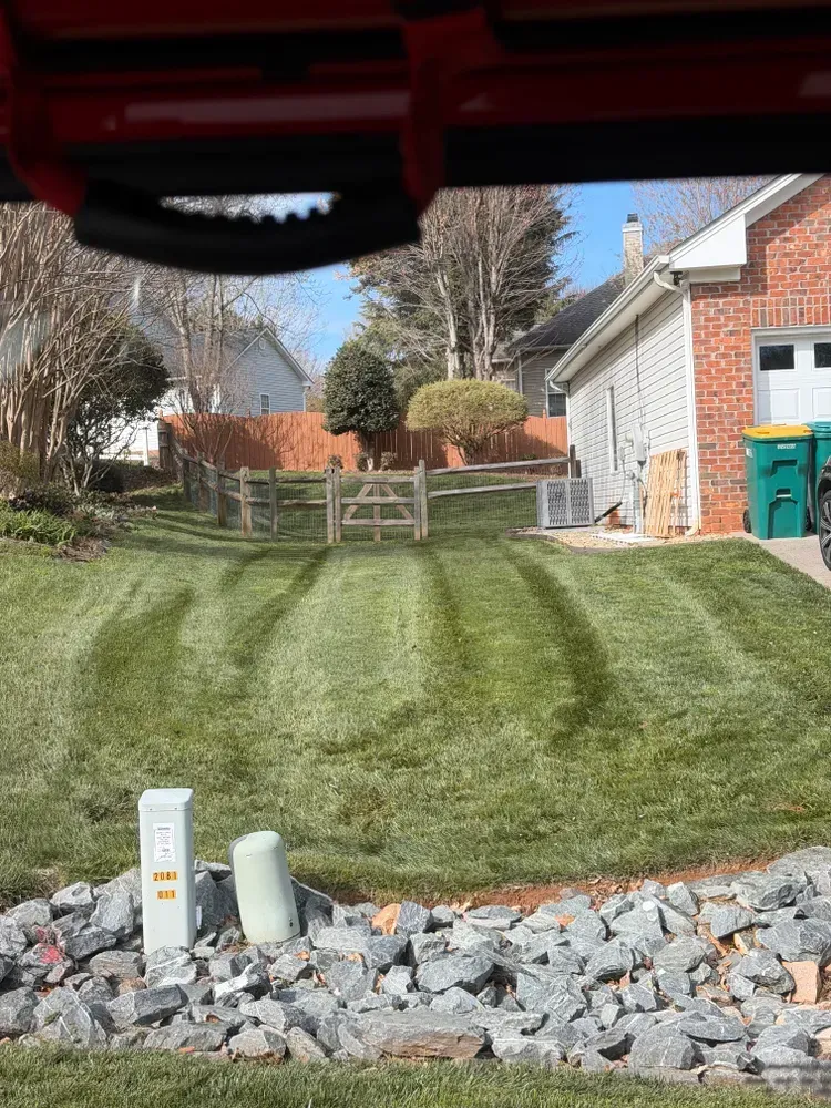 Backyard lawn with a wooden gate, stone border, and house beside a driveway.