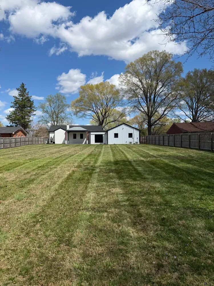 Wide mowed backyard leading to a white house and shed under a blue sky with clouds
