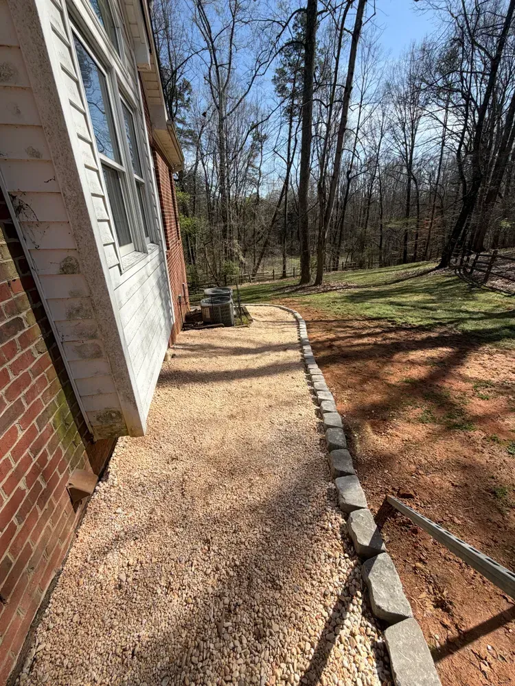 Side yard beside a brick house with a gravel path, stone border, and wooded backyard.
