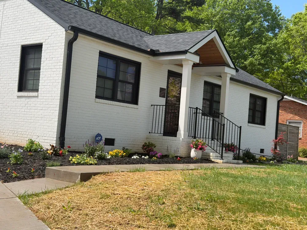 White brick house with black trim, small front porch, flower beds, and a sloped lawn under trees
