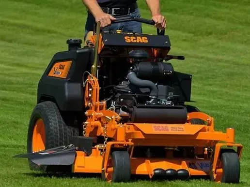 Orange SCAG riding lawn mower being driven on grass by a person in a black shirt.