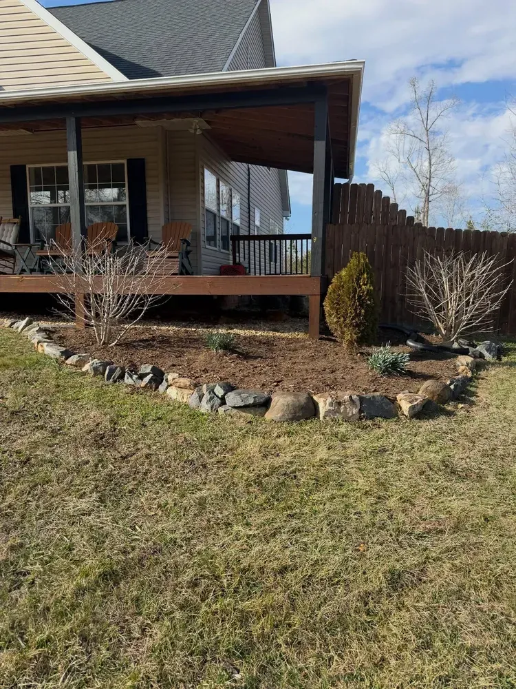 Front porch of a house with a rock-edged garden bed and leafless shrubs in a grassy yard.
