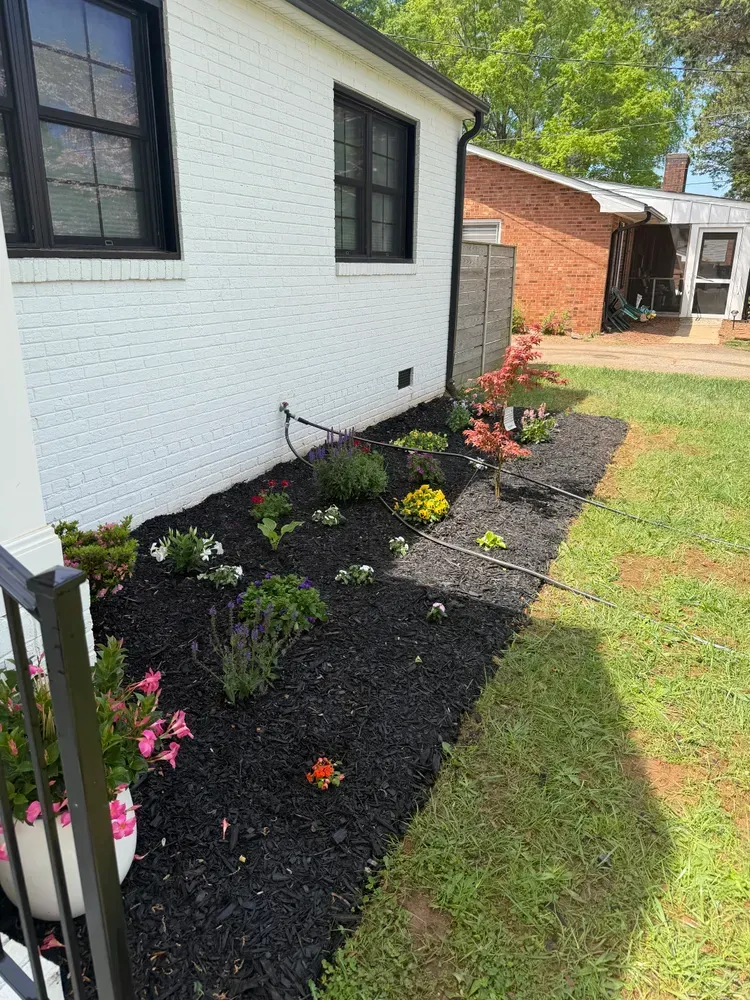 Small landscaped flower bed with colorful plants along a white house beside a grassy yard