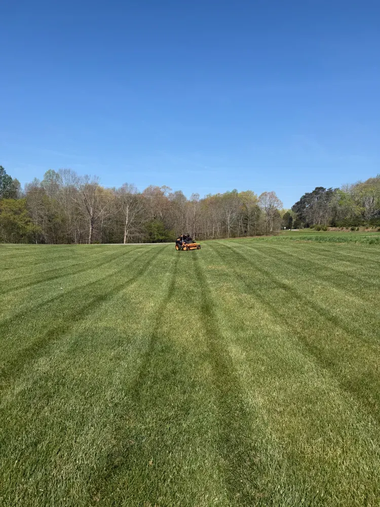 Wide green field with tire tracks, a small red tractor in the distance, and trees under a blue sky