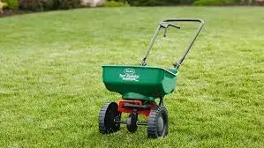 Green push spreader on a grassy lawn with black wheels and a metal handle.