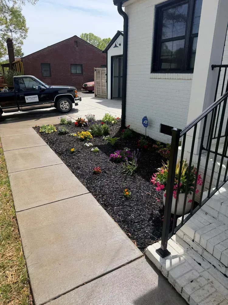 Front walkway beside white house with black railing and flower bed of colorful blooms; pickup truck in driveway.